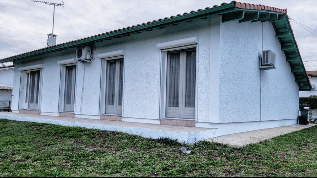 Façade de maison individuelle blanche, toit tuiles rouges, gouttière verte, volets roulants gris, deux climatisations et jardin en herbe.