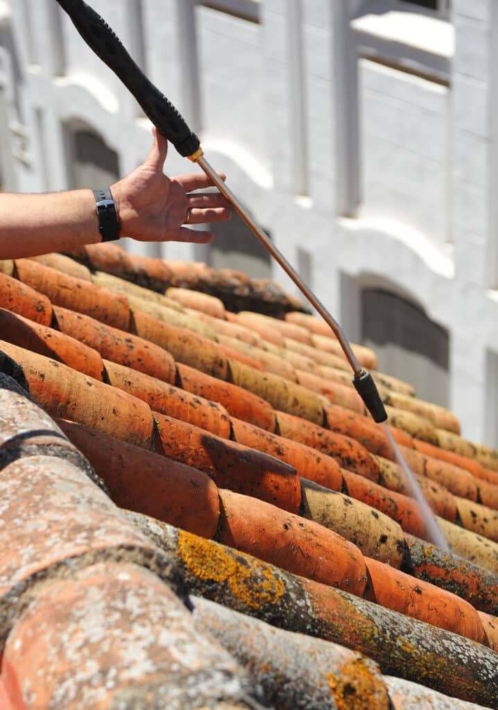 Main utilisant un nettoyeur haute pression pour nettoyer un toit de tuiles en terre cuite couvertes de mousse et lichen.