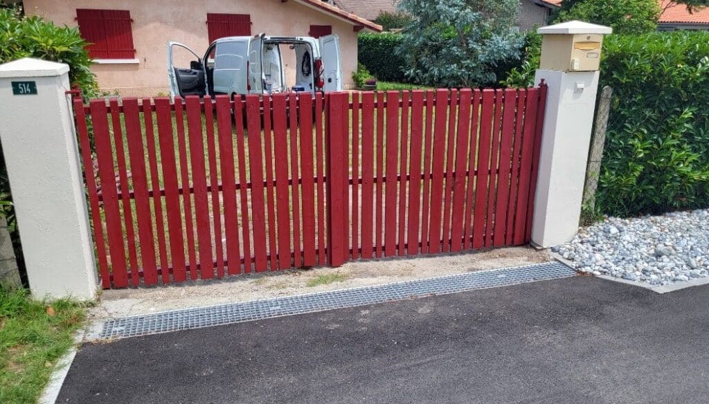 Portail Bois Rouge 514: Entrée Maison Moderne et Fonctionnelle Portail d'entrée rouge en bois avec poteaux blancs, numéro 514 et boîte aux lettres. Allée asphaltée, caniveau métallique. Van garé devant la maison.