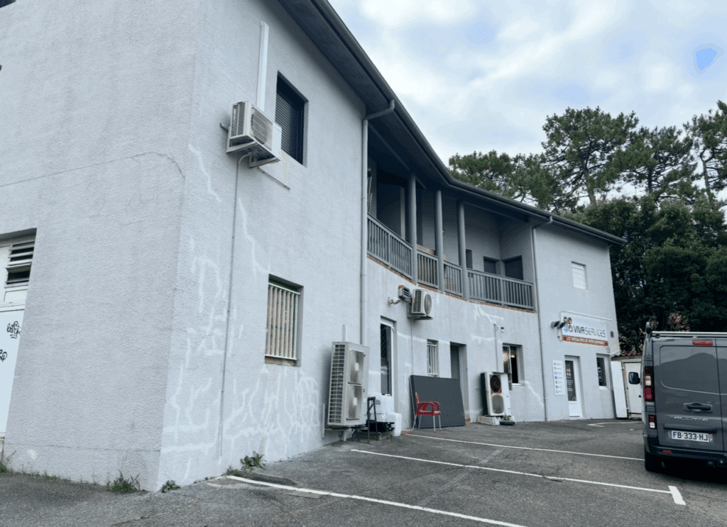 VIVA SERVICES : Bâtiment moderne, climatisation et parking Bâtiment gris clair de VIVA SERVICES avec plusieurs unités de climatisation, façade craquelée et un van garé sur le parking.