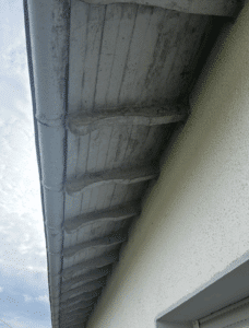 Vue en contre-plongée sous un débord de toit ancien avec gouttière grise et soffite en bois patiné, mur texturé clair.