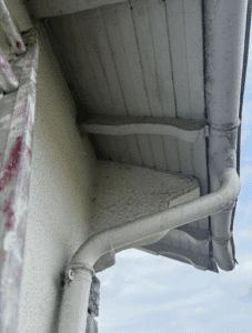 Détail sous un avant-toit avec gouttière et descente d'eau pluviale peintes en blanc, surplombant un ciel nuageux.