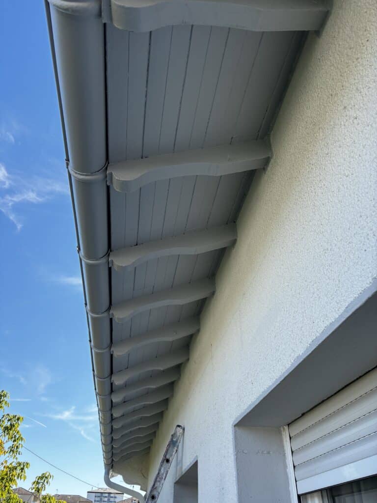 Ravalement de façade et peinture des sous-faces de toit gris clair, avec gouttière PVC et ciel bleu.